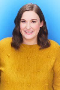A photograph of Chelsey B. Coombs wearing a yellow sweater in front of a blue background